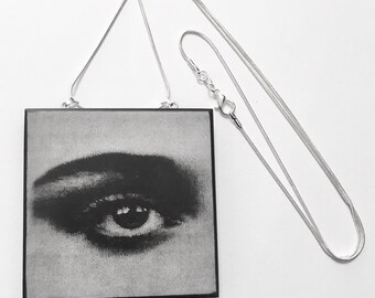 May include: A black and white photo of an eye, printed on a square wooden plaque. The plaque is attached to a silver chain necklace.