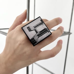 Può includere: Anello quadrato in bianco e nero con design architettonico. L'anello, indossato su un dito, mostra un'immagine dettagliata di un edificio. Il design crea un forte contrasto su uno sfondo neutro, creando un'estetica moderna.