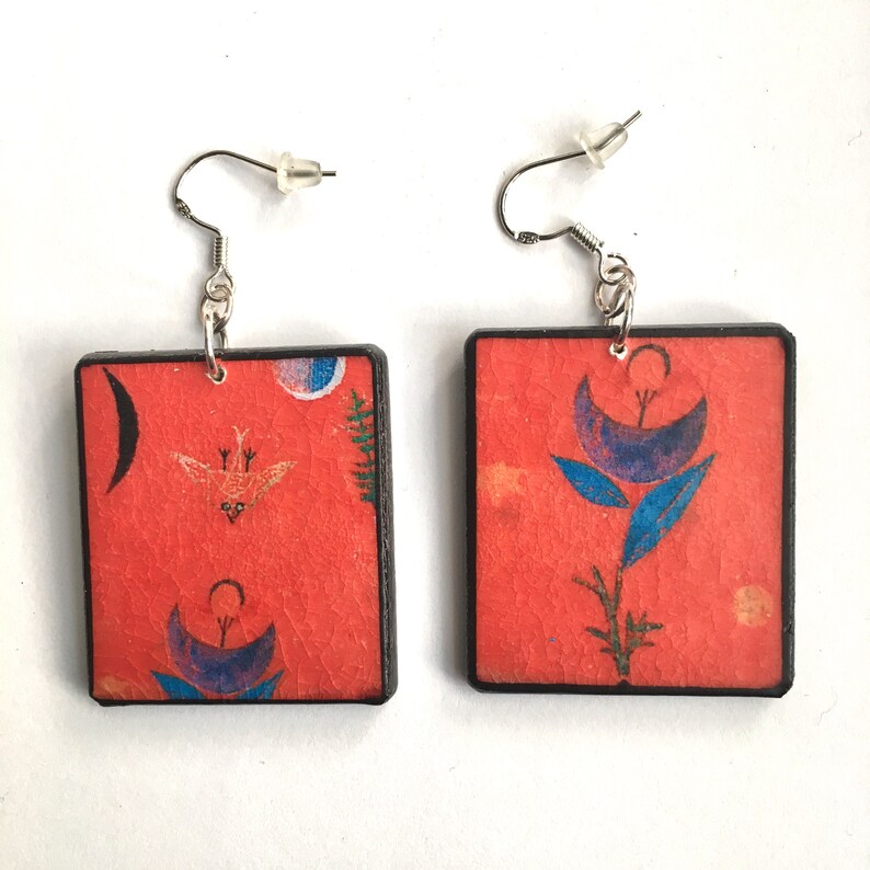 A pair of square earrings with a red background and black border. One earring features a blue flower with a yellow centre and the other earring features a bird with a yellow body and black wings.