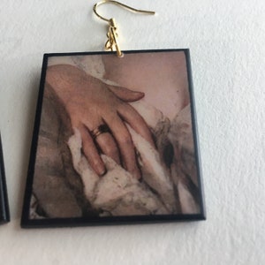 May include: Square earrings with a vintage image of a hand wearing a gold ring. The image is framed with a black border.