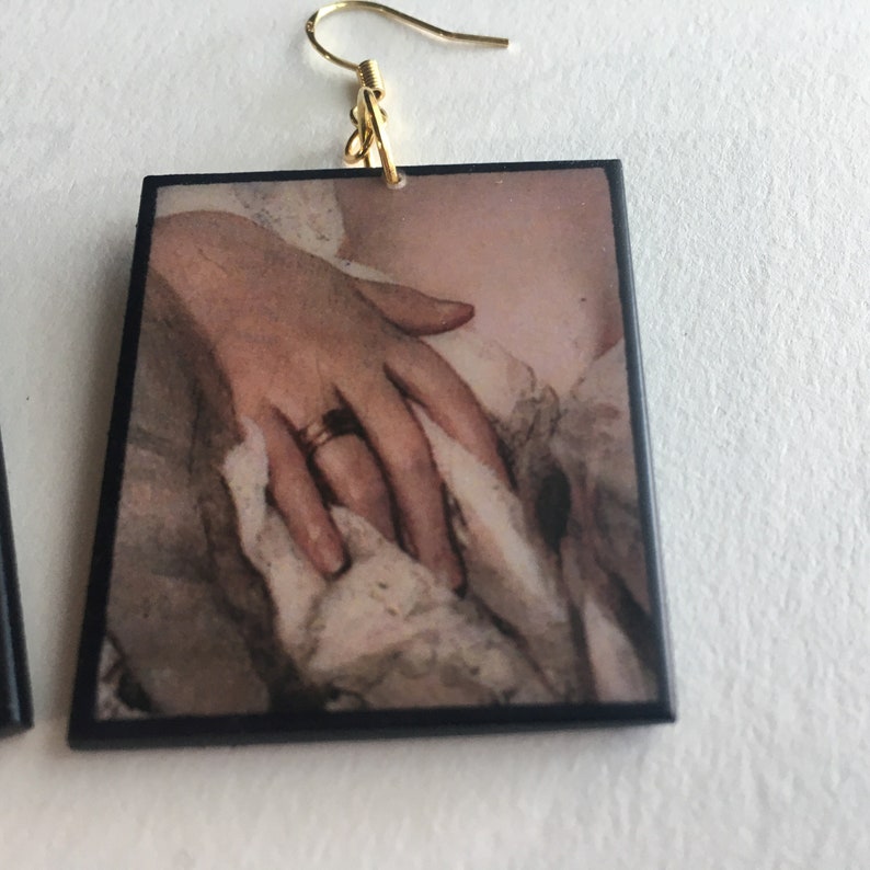 May include: Square earrings with a vintage image of a hand wearing a gold ring. The image is framed with a black border.