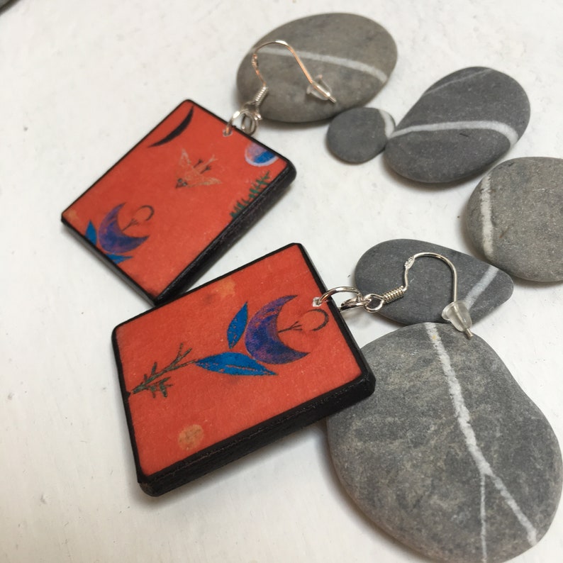May include: A pair of handmade earrings featuring square wooden tiles with a floral design and a blue and orange color scheme. The tiles are attached to silver ear wires and are accented with gray stones.