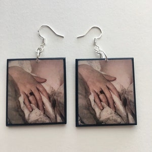 May include: A pair of square earrings with a black frame and a vintage image of a hand with a ring on it. The earrings have silver hooks.