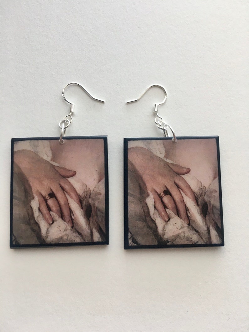 May include: A pair of square earrings with a black frame and a vintage image of a hand with a ring on it. The earrings have silver hooks.