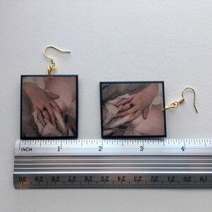 May include: A pair of earrings with black square-shaped pendants featuring a vintage photograph of a hand holding a piece of fabric. The earrings are made of gold-colored metal.