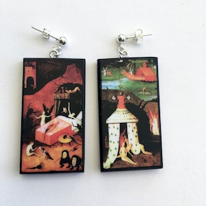 May include: A pair of black rectangular earrings with silver hooks. Each earring features a different scene from Hieronymus Bosch's 'The Garden of Earthly Delights' painting. The left earring shows a scene of hell with demons and a bed, while the right earring shows a scene of paradise with a white tent and figures.