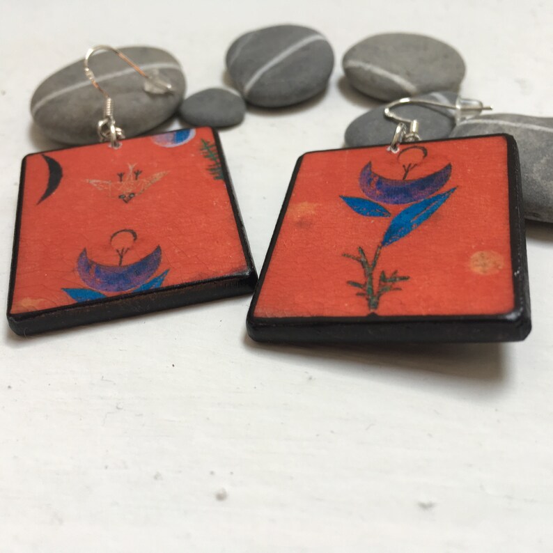 May include: A pair of square earrings with a red background and black border. The earrings feature a blue and purple floral design with a silver hook.