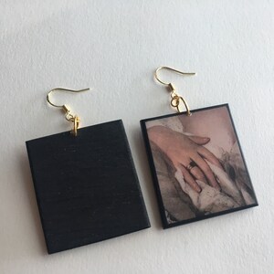 May include: A pair of square earrings. One earring is black with a gold hook. The other earring is a square picture of a hand with a ring on it. The picture is framed with a black border.