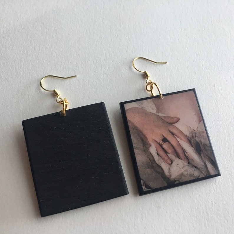 May include: A pair of square earrings. One earring is black with a gold hook. The other earring is a square picture of a hand with a ring on it. The picture is framed with a black border.