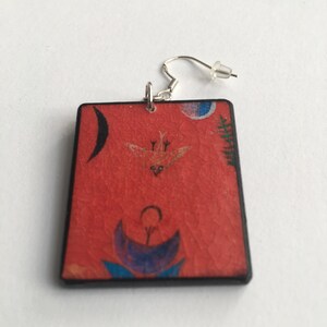 May include: Red rectangular earrings with a black border and a colorful abstract design featuring a bird, a crescent moon, and a flower.