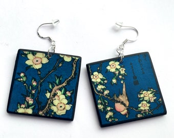 May include: A pair of square earrings with a blue background and a floral design featuring white flowers and a small bird. The earrings have silver hooks.