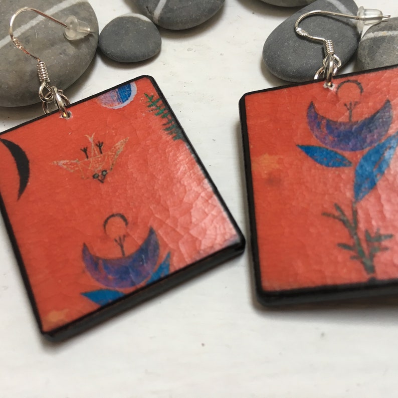 May include: A pair of square earrings with a red background and black trim. The earrings feature a floral design with blue and purple flowers and green leaves. One earring also features a small bird with yellow and orange accents.