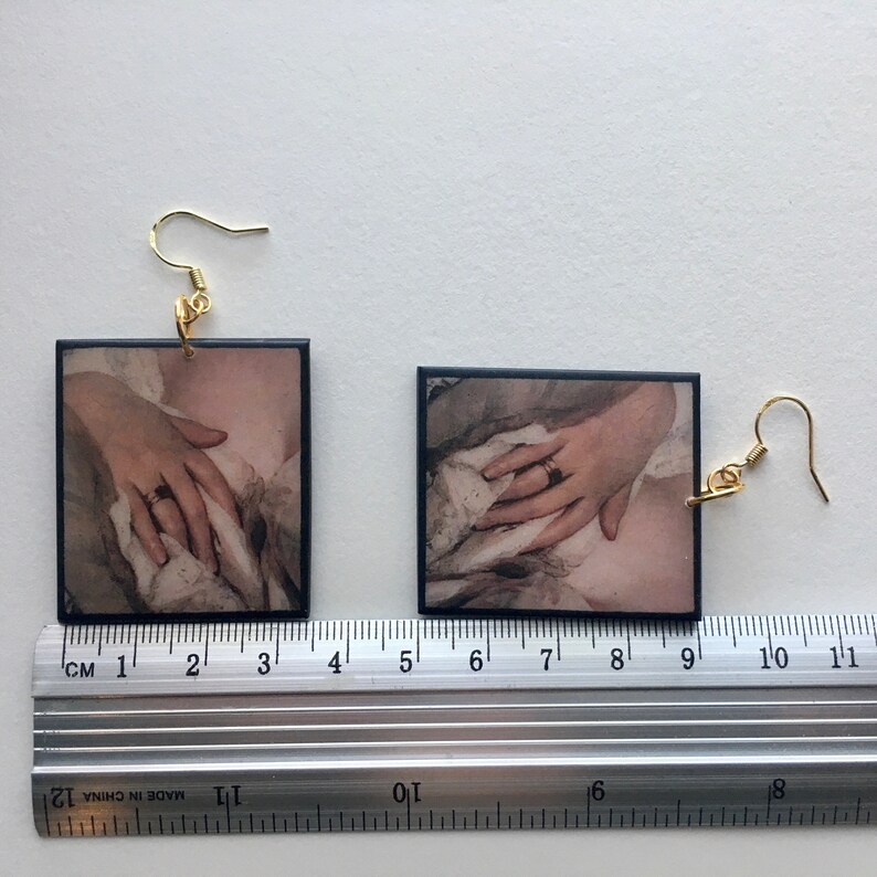 May include: A pair of earrings with black rectangular frames featuring a vintage style image of a hand with a ring. The earrings have gold hooks.