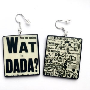 Op de afbeelding: Een paar vierkante oorbellen in zwart en wit met zilveren haakjes. De oorbellen bevatten een collage van tekst, waaronder de woorden "Wat is Dada?" op de ene oorbel en "Dada" op de andere.