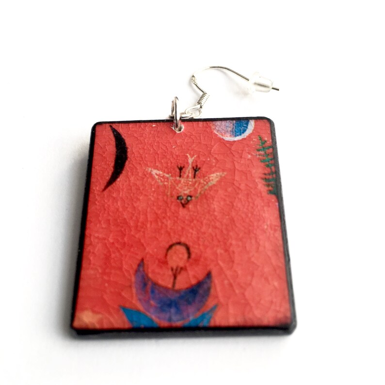 May include: Red rectangular earrings with a black border and a colorful abstract design featuring a bird, a crescent moon, and a flower.