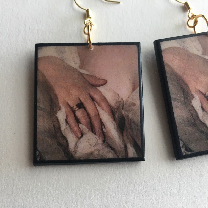 May include: A pair of square earrings with a vintage image of a hand wearing a ring. The earrings have gold hooks.