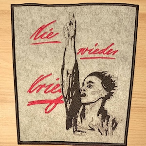 Könnte beinhalten: Ein grauer gestickter Aufnäher mit einem Schwarzweißbild einer Person, die die Faust in die Luft reckt. Der Text "Der Krieg" und "wieder" ist in Rot gestickt.