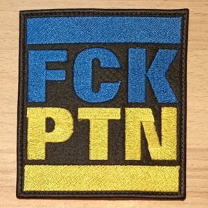 May include: A black embroidered patch with the text "FCK PTN" in blue and yellow thread. The patch features a blue and yellow stripe design, resembling the Ukrainian flag.