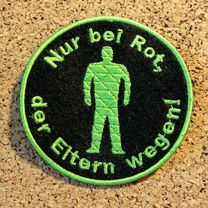 May include: A circular black patch with a green border. It features a green embroidered figure of a person and the text "Nur bei Rot, der Eltern wegen!" in green, encircling the figure.
