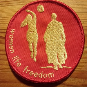 Peut inclure: Patch rond rouge avec des figures brodées en or de deux personnes jouant au volley-ball. Le texte "Women Life Freedom" est brodé en or autour du bord.