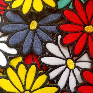 Può includere: Una collezione di toppe di fiori in feltro colorati in varie tonalità di rosso, giallo, blu e bianco. I fiori hanno un semplice design a margherita con un centro giallo e più petali.