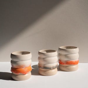 Könnte beinhalten: Drei Keramikgefäße mit zylindrischer Form und strukturiertem, geschichtetem Design. Die Gefäße sind überwiegend cremefarben mit orangefarbenen und beigefarbenen Bändern. Die Objekte werden auf einer weißen Oberfläche präsentiert.