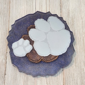 May include: A resin coaster with a dark blue, glittery background and a wavy edge. It features three paw prints: two white and one brown. The coaster is designed to protect surfaces from drink rings.