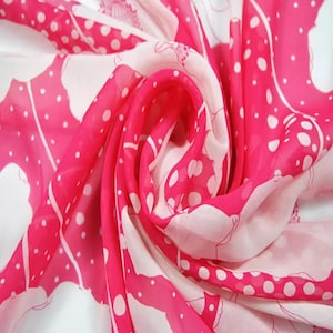 Może przedstawiać: Zbliżenie różowo-białego szalika z kwiatowym i groszkowym wzorem. Szalik wykonany jest z lekkiej, zwiewnej tkaniny. Wzór przedstawia duże, stylizowane kwiaty i małe białe grochy na jasnoróżowym tle.