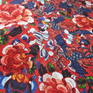 Pode incluir: Um tecido vibrante com um padrão floral e de pássaros repetido. O design apresenta grandes rosas vermelhas, pássaros azuis e detalhes florais brancos e azuis sobre um fundo vermelho. O tecido tem uma aparência detalhada e texturizada.