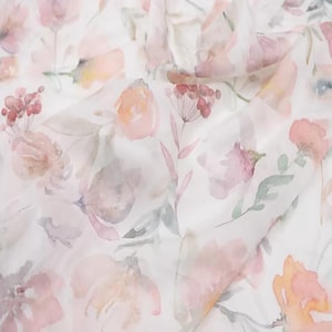 Op de afbeelding: Een close-up van een stof met een aquarel bloemenpatroon. Het ontwerp toont zachte roze, perzik en gedempte groene bloemen en bladeren op een witte achtergrond. De stof heeft een delicate, doorschijnende uitstraling.