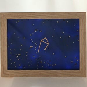 以下が含まれることがあります： 暗い青色の背景に金色のグリッターの星が散りばめられた、金色のワイヤーで描かれた天秤座の星座を表現した額縁付きの星座アート作品です。