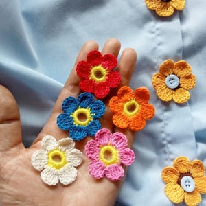 Puede incluir: Una colección de aplicaciones de flores de ganchillo de colores, incluyendo rojo, azul, naranja, rosa, blanco y amarillo. Algunas flores están unidas a una tela azul claro con botones. Las flores tienen centros amarillos.