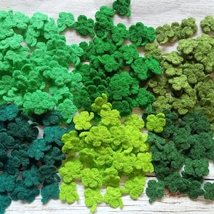 Può includere: Una collezione di trifogli all'uncinetto in varie tonalità di verde, dal verde lime chiaro al verde foresta scuro. Le foglie di trifoglio fatte a mano sono sparse su una superficie di legno, perfette per lavori artigianali e decorazioni per San Patrizio.