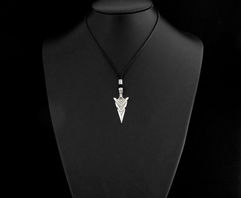 Puede incluir: Un collar con un colgante de plata con un dise&ntilde;o de punta de flecha geom&eacute;trica. El colgante est&aacute; en un cord&oacute;n negro.