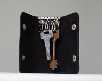 May include: Black leather key holder with silver metal key rings. Two keys are attached, one silver and one bronze. The key holder is open, revealing the key rings and keys. The key holder is secured with snap closures.