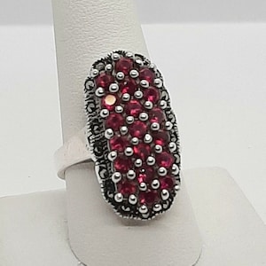 May include: A silver ring with a large oval-shaped red gemstone surrounded by smaller silver stones.