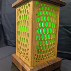 May include: A rectangular wooden lamp with a dark brown base and top. The sides are made of light brown bamboo with a pattern of oval cutouts. Bright green light shines through the cutouts, illuminating the interior.
