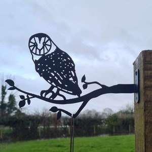 May include: Black metal owl silhouette perched on a branch with a bird feeder hanging below. The owl and branch are mounted to a wooden post.