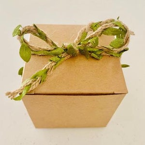 Könnte beinhalten: Eine braune Geschenkbox aus Pappe mit einer grünen Schleife aus Bast und Blättern. Die Box ist mit Bast gebunden und mit grünen Blättern verziert.