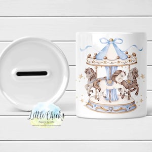 May include: White ceramic money box with a slot on the top. The front of the box features a blue and white carousel with brown horses. The text "Little Chickie Paperie & Gifts" is printed on the bottom of the box.