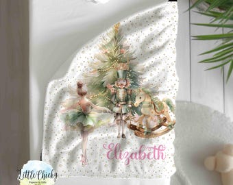 Könnte beinhalten: Eine weiße Babydecke mit einem goldenen und weißen Schneeflockenmuster. Die Decke zeigt eine Aquarell-Illustration einer Ballerina, eines Nussknackers, eines Schaukelpferdes und eines Weihnachtsbaums. Der Name "Elizabeth" ist in rosa kursiver Schrift geschrieben.