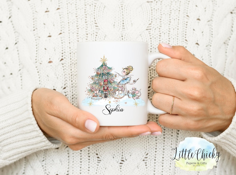 May include: White ceramic mug with a Christmas-themed design featuring a ballerina, a nutcracker, a mouse, and a Christmas tree. The mug has the name "Sophia" written on it.