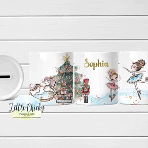 Könnte beinhalten: Weiße Keramik-Spardose mit Schlitz und drei passende Becher. Die Becher zeigen Illustrationen eines Weihnachtsbaums, eines Schaukelpferds, eines Nussknackers und Ballerinas. Ein Becher ist mit dem Namen "Sophia" in goldfarbenen Buchstaben personalisiert.