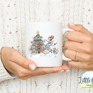 May include: White ceramic mug with a colorful illustration of a ballerina decorating a Christmas tree with a nutcracker and mice. The mug has a gold handle and a gold rim.