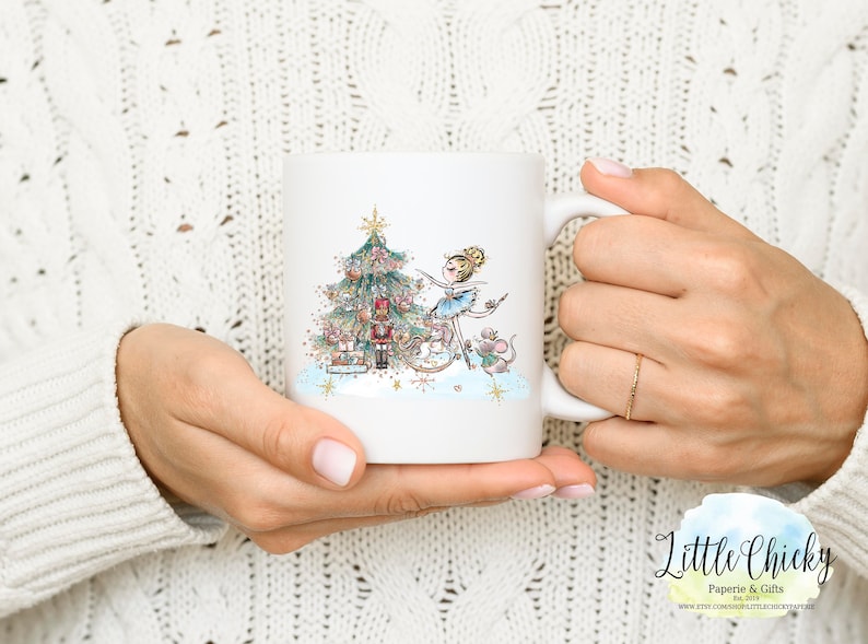 May include: White ceramic mug with a colorful illustration of a ballerina decorating a Christmas tree with a nutcracker and mice. The mug has a gold handle and a gold rim.