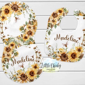 Op de afbeelding: Een set van twee baby-slabbetjes met een zonnebloem- en bijendesign. De slabbetjes zijn wit met een bruine rand en bevatten de naam "Madeline" in bruine tekst. De slabbetjes zijn gemaakt door Little Chickie Paperie & Gifts.