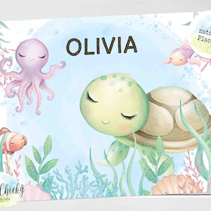Puede incluir: Una ilustración acuarela azul y verde de una tortuga, un pulpo y un pez payaso con el nombre Olivia. La ilustración está sobre un fondo blanco con el texto "A3 matching Placemat" en la esquina superior derecha.