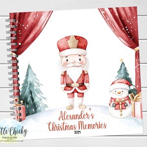 Könnte beinhalten: Ein rotes und weißes Weihnachts-Erinnerungsbuch mit einer Aquarell-Illustration eines Nussknackers und eines Schneemanns. Das Buch hat eine Spiralbindung und den Text "Alexander's Christmas Memories 2021" auf dem Cover.
