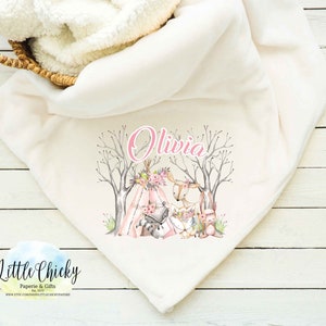 May include: A white fleece baby blanket with a pink and brown woodland animal design. The blanket features a deer, a fox, and a raccoon sitting around a teepee. The name "Olivia" is written in pink above the animals.