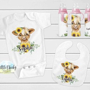 Pode incluir: Conjunto para bebé com body branco, babete e biberões. Todos apresentam uma ilustração de uma vaca das Terras Altas com girassóis e o nome "Sophie". Os biberões têm detalhes cor-de-rosa.